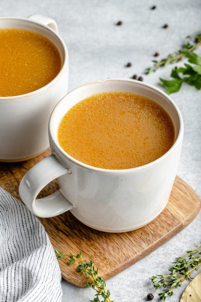 Deux tasses de bouillon d'os matinal fumant, posées sur une planche en bois avec des herbes aromatiques pour la routine bien-être Morning Broth.